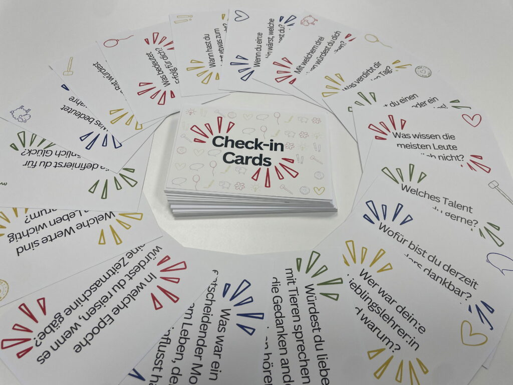 Check-in Cards mit farbigen Symbolen und Gesprächsfragen, aufgefächert um einen Kartenstapel mit Logo.